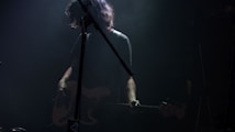 A musician stands in subdued lighting with a guitar, partially obscured by shadows and stage equipment. The scene conveys a moody, intimate atmosphere often associated with live music performances.