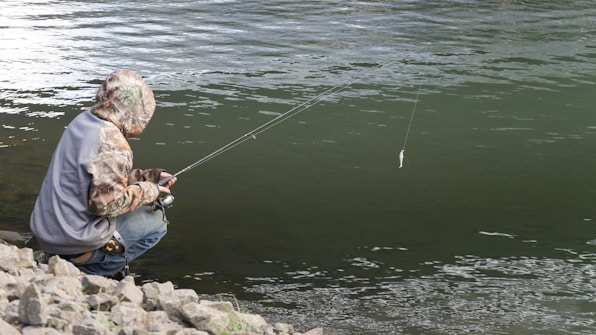 A person crouches on rocky terrain beside a body of water, holding a fishing rod with a line extended over the water. The individual is wearing a camouflage jacket and a gray vest, appearing focused on fishing.