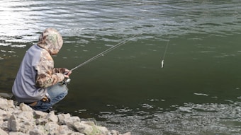 A person crouches on rocky terrain beside a body of water, holding a fishing rod with a line extended over the water. The individual is wearing a camouflage jacket and a gray vest, appearing focused on fishing.