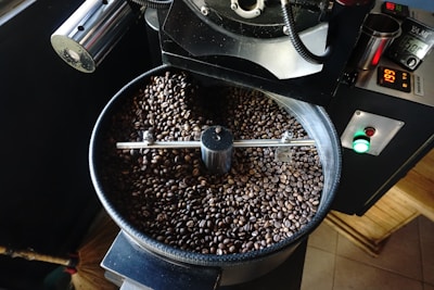 A coffee roasting machine is filled with roasted coffee beans. The machine appears to be in mid-operation, with a digital display showing the temperature. The beans are dark and glossy, suggesting they are freshly roasted.