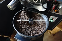 A coffee roasting machine is filled with roasted coffee beans. The machine appears to be in mid-operation, with a digital display showing the temperature. The beans are dark and glossy, suggesting they are freshly roasted.