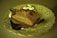 A golden brown pastry rests on a piece of foil, placed on an elegant, ornate white plate with a scalloped edge and delicate cut-out patterns. The lighting casts a warm, yellowish tint over the entire scene, highlighting the crisp texture of the pastry.