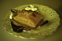 A golden brown pastry rests on a piece of foil, placed on an elegant, ornate white plate with a scalloped edge and delicate cut-out patterns. The lighting casts a warm, yellowish tint over the entire scene, highlighting the crisp texture of the pastry.