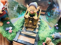 A whimsical snow globe features a playful character riding in a small wooden cart on a checkered path surrounded by lush green grass and colorful flowers. The scene includes two adorable animals, resembling a bear and a dog, adding to the charm. Glittering particles inside the globe create a magical atmosphere.
