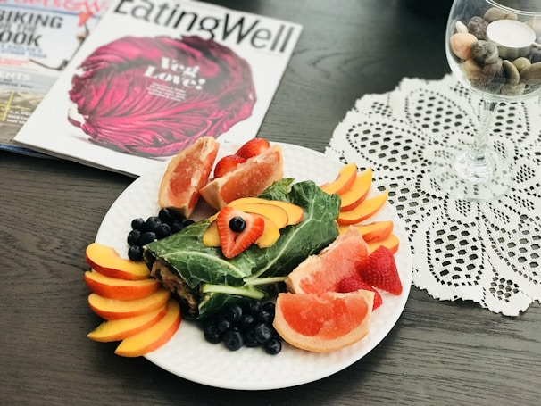 A decorative plate with a variety of sliced fruits like grapefruit, peach, strawberry, and blueberries arranged around a wrapped leafy vegetable centerpiece. There are magazines titled 'EatingWell' and others in the background. To the right is a glass with decorative pebbles placed on a lace tablecloth.