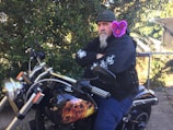 A man sits confidently on a motorcycle with arms crossed, wearing a black jacket adorned with white flame graphics and a green beanie. A plush toy with bright pink fur and large eyes is perched on his shoulder, adding a whimsical touch. The motorcycle features artwork of a flaming skull on the fuel tank. The background is rich with green foliage, and sunlight filters through the leaves.