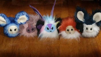 Five plush toys with large eyes and various colors are arranged in a line. Each toy has distinct features: the first is predominantly blue with large ears, the second is pink with a horn resembling a unicorn, the middle toy is colorful with a mix of blue, purple, and yellow, the fourth toy is orange with a mischievous expression, and the last one is similar to the first but with darker blue and furry ears.