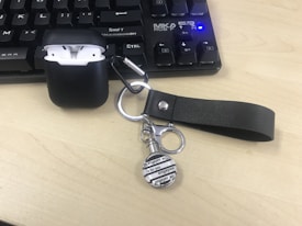 A pair of white wireless earbuds are in a black protective case. Attached to the case is a keychain with a metal clasp and a decorative element inscribed with text. The items rest on a wooden or laminate surface next to a black computer keyboard with illuminated keys.