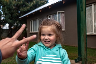 A young child wearing a turquoise hoodie with white stripes smiles while holding up a peace sign. In the foreground, a hand also makes a peace sign gesture. The scene takes place in an outdoor setting, with a house and green grass in the background.
