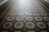 An ornate tiled floor with a hexagonal pattern, featuring intricate designs in shades of blue and beige. The central motif of each tile includes floral elements outlined in different colors.