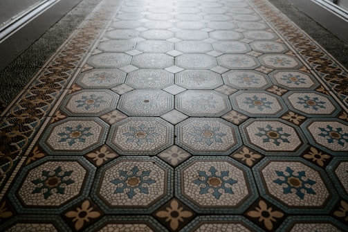 An ornate tiled floor with a hexagonal pattern, featuring intricate designs in shades of blue and beige. The central motif of each tile includes floral elements outlined in different colors.