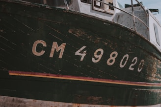 A close-up shot reveals the weathered hull of a boat, marked with the registration number CM 498020. The paint appears chipped and worn, indicating age and exposure to the elements. Wooden parts of the boat's structure are visible, adding to its rugged and rustic appearance.
