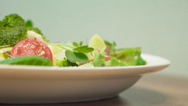 A close-up of a fresh salad on a white plate. The salad contains colorful vegetables, including a slice of tomato, broccoli florets, and various leafy greens. The background is softly blurred, highlighting the vibrant colors and textures of the salad.