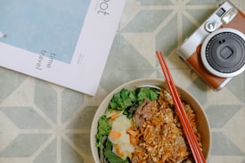 A bowl filled with a mixture of greens, pickled vegetables, meat topped with crispy fried pieces, and a pair of red chopsticks rests on it. Nearby, on a geometric patterned surface, a vintage-style camera and a magazine labeled 'time travel' are placed.