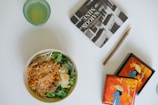 A bowl of salad with shredded carrots, lettuce, and meat, next to a glass with a light green drink. There is a book titled 'The Saigon of Mine' and a rustic pencil, along with a decorative box featuring an illustration of a person in a traditional hat against a bright orange background with floral designs.