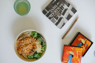 A bowl of salad with shredded carrots, lettuce, and meat, next to a glass with a light green drink. There is a book titled 'The Saigon of Mine' and a rustic pencil, along with a decorative box featuring an illustration of a person in a traditional hat against a bright orange background with floral designs.