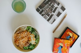 A bowl of salad with shredded carrots, lettuce, and meat, next to a glass with a light green drink. There is a book titled 'The Saigon of Mine' and a rustic pencil, along with a decorative box featuring an illustration of a person in a traditional hat against a bright orange background with floral designs.