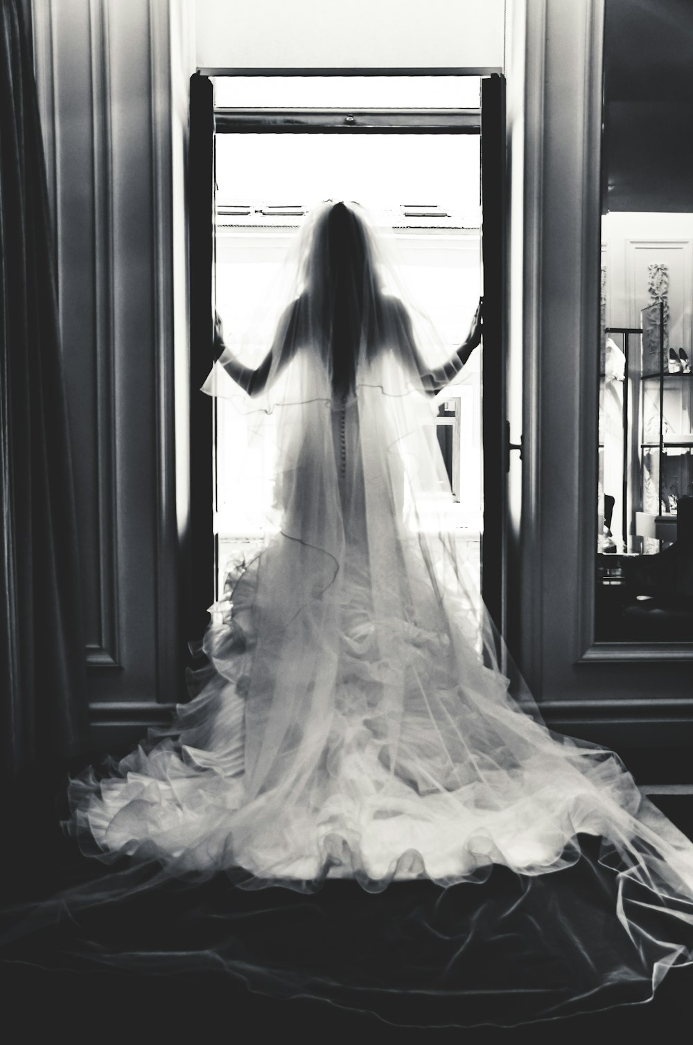 grayscale photography of woman wearing wedding gown standing near open window