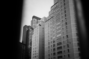 A black and white photograph capturing a cluster of tall, residential buildings. The structures are modern, with numerous windows arranged in a grid pattern. The angle of the photo gives a sense of height and urban density.