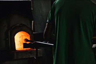 A person stands in front of a glowing furnace, using a long metal rod to interact with the intense heat inside. The ambient lighting is dim, highlighting the vibrant orange and yellow hues emanating from the furnace's opening.