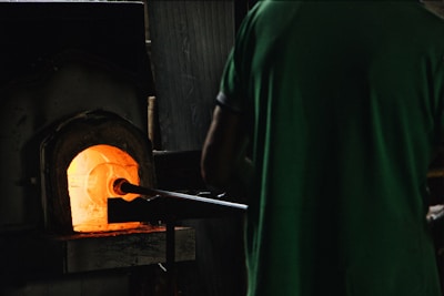 A person stands in front of a glowing furnace, using a long metal rod to interact with the intense heat inside. The ambient lighting is dim, highlighting the vibrant orange and yellow hues emanating from the furnace's opening.