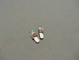 A pair of multi-colored sandals lies on a smooth, flat sandy surface. The sandals have vibrant accents of blue and brown, contrasting with the muted sand.