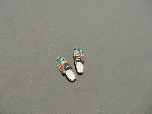 A pair of multi-colored sandals lies on a smooth, flat sandy surface. The sandals have vibrant accents of blue and brown, contrasting with the muted sand.