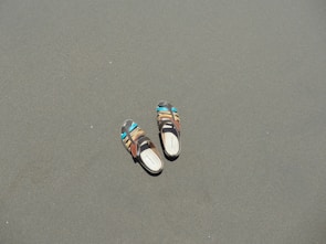 A pair of multi-colored sandals lies on a smooth, flat sandy surface. The sandals have vibrant accents of blue and brown, contrasting with the muted sand.
