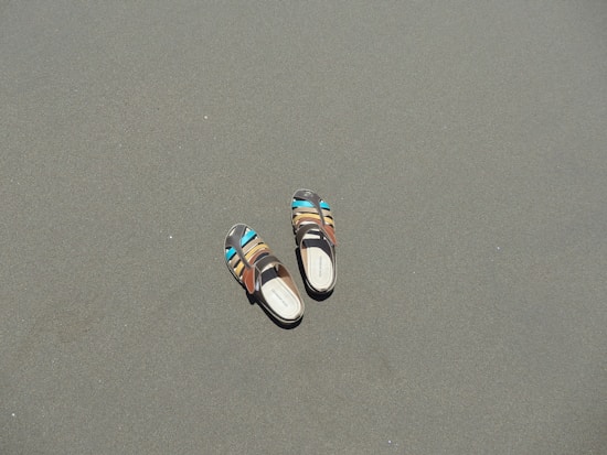 A pair of multi-colored sandals lies on a smooth, flat sandy surface. The sandals have vibrant accents of blue and brown, contrasting with the muted sand.