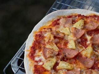 A freshly baked pizza sits on a metal grill, topped with slices of ham and chunks of pineapple over a layer of marinara sauce and melted cheese. The crust appears thin and crispy, with a slightly browned texture.