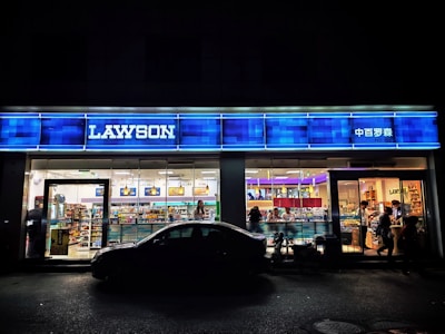 A convenience store with a brightly lit interior and a blue sign displaying 'LAWSON'. The storefront is filled with various products and several people are visible inside shopping. A car is parked in front, creating a silhouette against the illuminated glass windows.