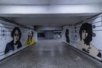 A dimly lit underground passageway featuring large murals on both walls. On the left, a mural depicts a person in dark glasses, next to a bright yellow image of someone singing into a microphone. On the right wall, there are graphic-style portraits including text and musical notes.