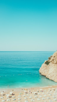 A scenic beach view with clear turquoise water and a rocky cliff to the right. Numerous beach umbrellas and chairs line the sandy shore, with people enjoying the sun and sea. The sky is a bright, clear blue, enhancing the vacation-like scenery.