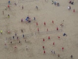 Aerial view of a crowded beach volleyball area with multiple games happening simultaneously. People in colorful athletic wear are actively engaged in playing or watching. The sandy expanse is marked with volleyball court lines.