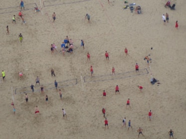 Aerial view of a crowded beach volleyball area with multiple games happening simultaneously. People in colorful athletic wear are actively engaged in playing or watching. The sandy expanse is marked with volleyball court lines.