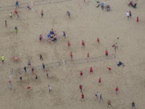 Aerial view of a crowded beach volleyball area with multiple games happening simultaneously. People in colorful athletic wear are actively engaged in playing or watching. The sandy expanse is marked with volleyball court lines.