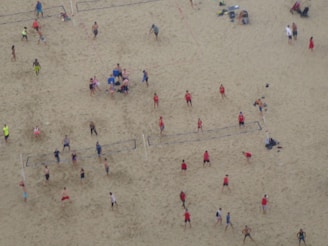 Aerial view of a crowded beach volleyball area with multiple games happening simultaneously. People in colorful athletic wear are actively engaged in playing or watching. The sandy expanse is marked with volleyball court lines.