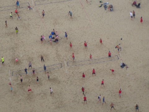 Aerial view of a crowded beach volleyball area with multiple games happening simultaneously. People in colorful athletic wear are actively engaged in playing or watching. The sandy expanse is marked with volleyball court lines.