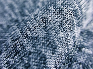 Close-up view of a textured fabric surface with intricate stitching and a wavy pattern. The threads appear in various shades of blue, giving depth and dimension.