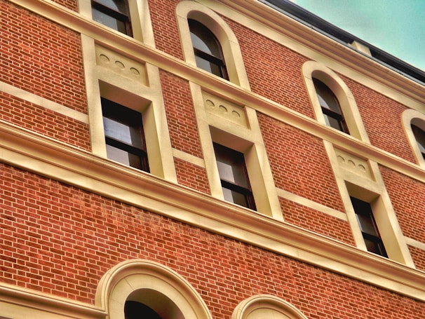 A brick facade of a building with multiple windows framed in a cream-colored trim. The brickwork is detailed and uniform, and the windows are evenly spaced with arch designs above them.