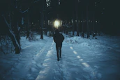 A shadowed figure walking a lonely path through a dark forest, light breaking through the trees ahead.