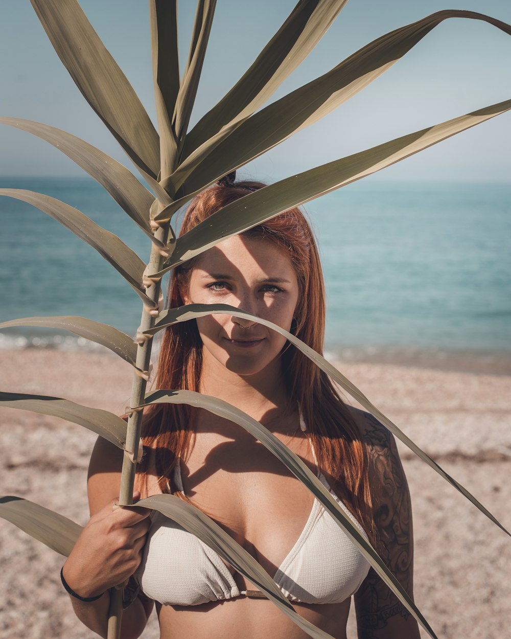 ビーチで顔の近くに線形の葉を保持している白いビキニトップの女性