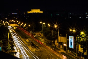 A nighttime cityscape with a brightly illuminated road and cars creating streaks of white and red light. Streetlights line the road, casting a warm glow. A large building in the background is prominently lit, and there's a billboard on the right side displaying text.