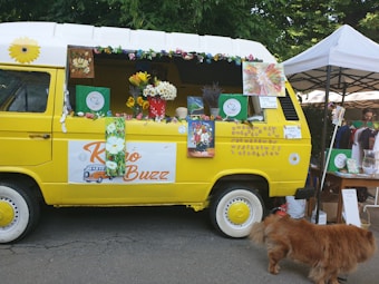 A bright yellow van decorated with colorful flowers and artwork, parked near a tent at an outdoor market. The side window of the van is open, displaying various items, including potted flowers and signs. A fluffy brown dog is walking nearby.