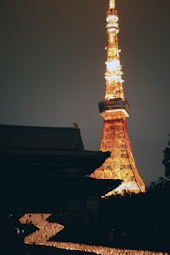 A tall, illuminated tower, possibly the Tokyo Tower, glowing brightly with warm orange and yellow lights against a dark sky. In the foreground, rows of lit candles form a winding path leading towards the tower, creating a serene and peaceful atmosphere. The silhouette of a traditional building, likely a temple, is visible beside the tower.