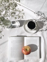 A sunlit café table with open books, a laptop, and a pastel-colored journal surrounded by pressed flowers.