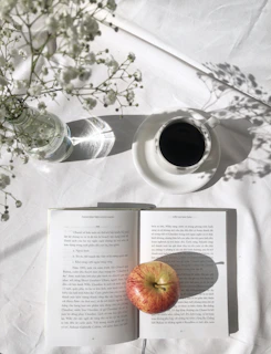 A sunlit café table with open books, a laptop, and a pastel-colored journal surrounded by pressed flowers.