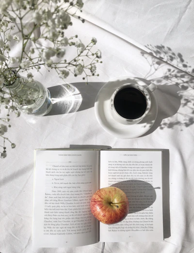 apple fruit on top of open book beside black coffee in white ceramic cup on table