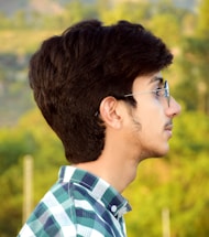 A young man with dark hair and glasses is seen in profile, wearing a plaid shirt. The background is blurred with greenery, suggesting an outdoor setting.