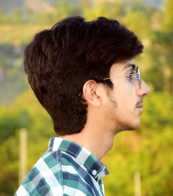A young man with dark hair and glasses is seen in profile, wearing a plaid shirt. The background is blurred with greenery, suggesting an outdoor setting.